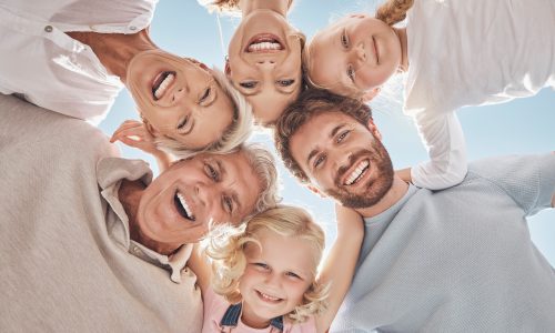 Happy family, huddle and smile below in trust, community or support together against a blue sky. Portrait of grandparents, parents and children hugging, smiling or bonding for holiday break in nature